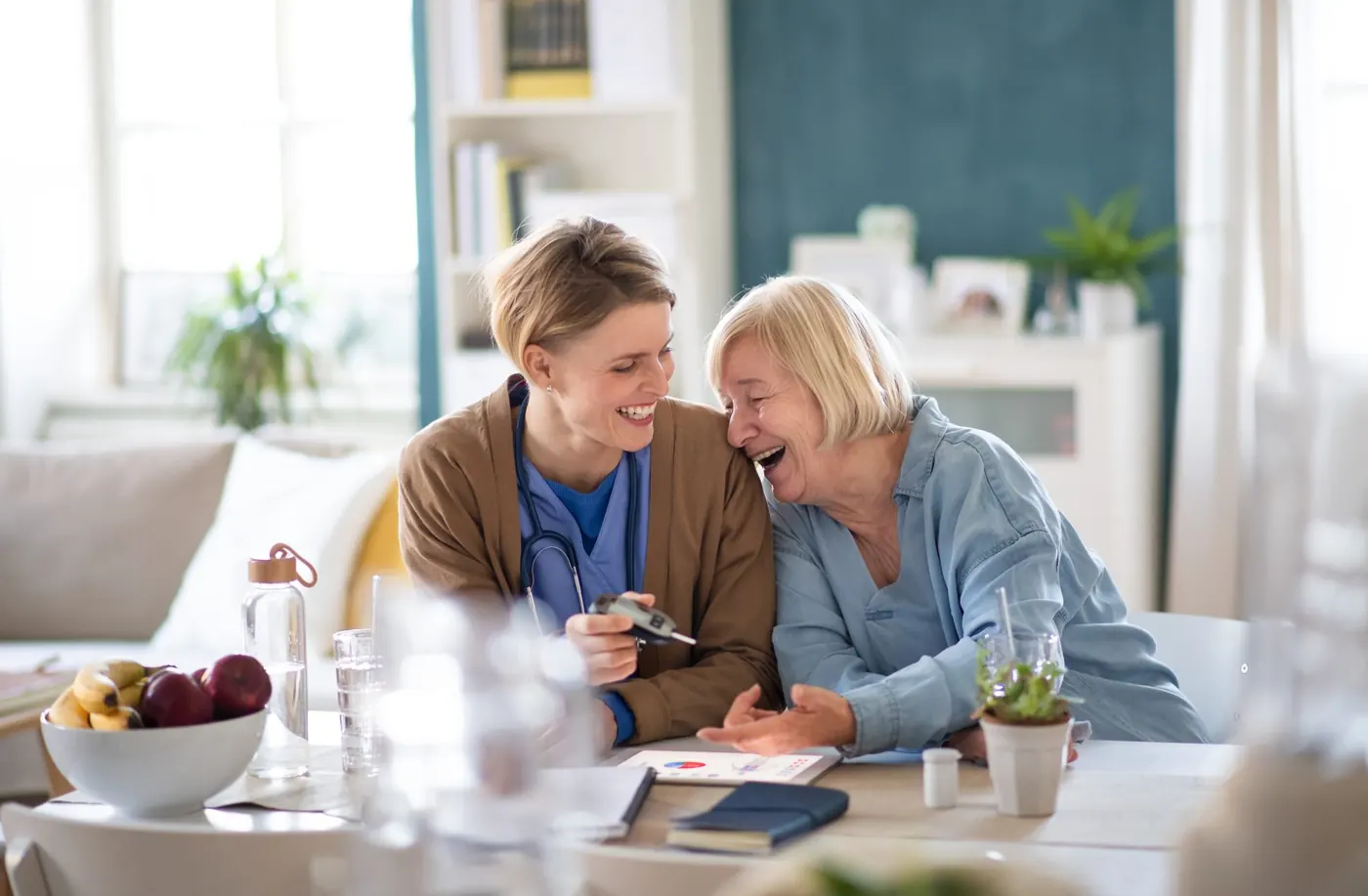 Una cuidadora y una señora mayor se ríen juntas sentadas a la mesa de la cocina.